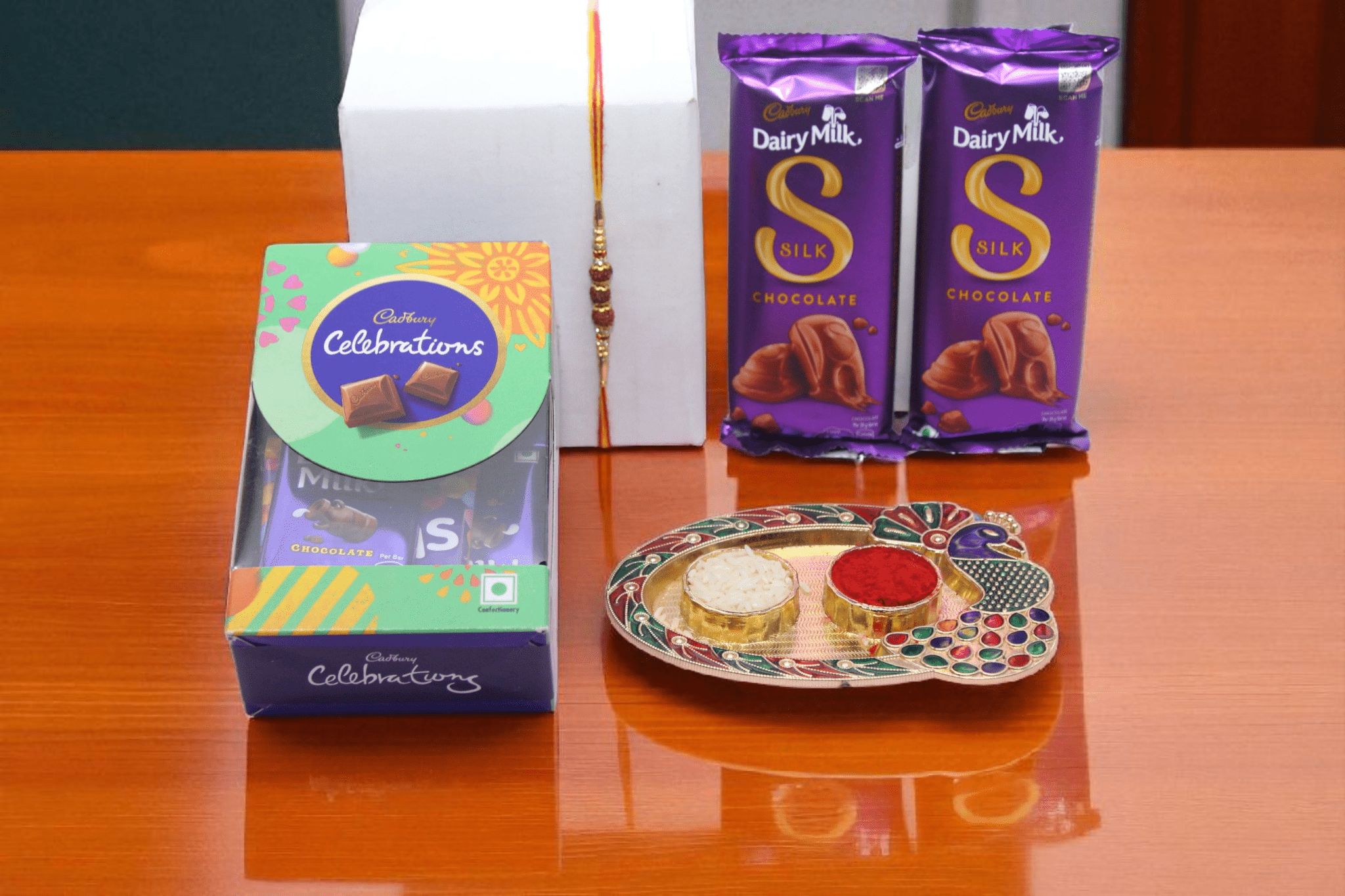 Rudraksha Chocolate Celebration Hamper. Rudraksha Chocolate Celebration Hamper with Rudraksha Rakhi, Cadbury Dairy Milk Silk Bars, Cadbury Celebrations Pack and Roli Chawal