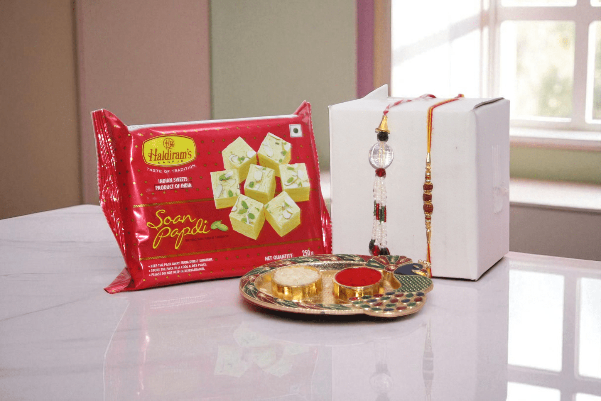 Rakhi With Sweet Bhaiya Bhabhi Rakhi gift combo featuring a designer Lumba Rakhi set, Haldiram Soan Papdi sweets, festive pooja thali with roli chawal, and white gift box – perfect Raksha Bandhan hamper for brother and sister-in-law. - YHOJA.COM